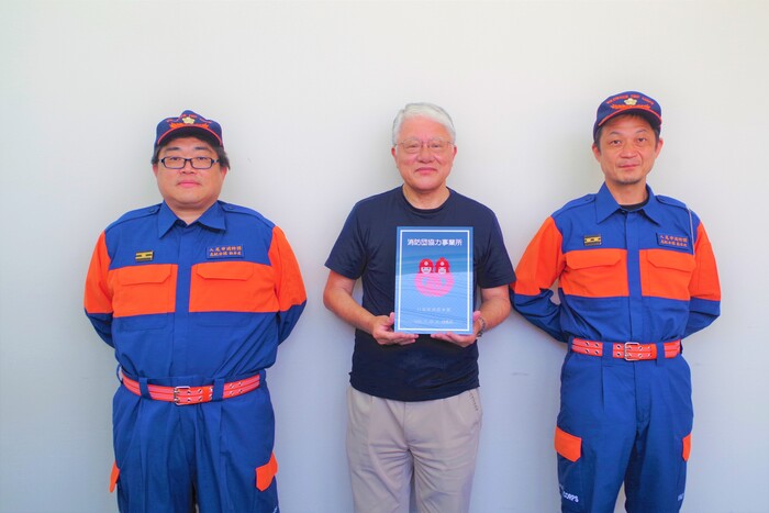 3名の写真が写っており左から松本団員中央が消防団協力事業所の標示証を持っている施設長永原さん右端が森本団員