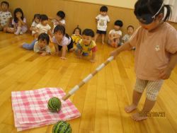 目隠しをして棒をもち、育てた小さいスイカの前に立つ子どもの写真