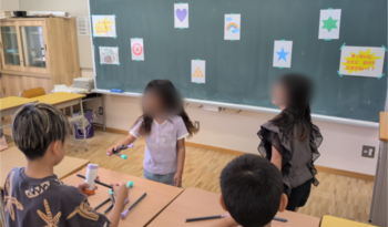 お店の説明を聞く子どもたち。黒板に、星や虹など様々な模様の的が貼られており、子どもたちは手作りの道具を使用して的を狙う。